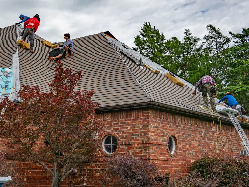 A roofing crew of six re-roofing a brick house, ladders up and bundles staged on the roof