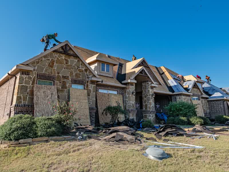 A crew tearing the old shingles off a stone and brick house, scrap shingles piled on the lawn